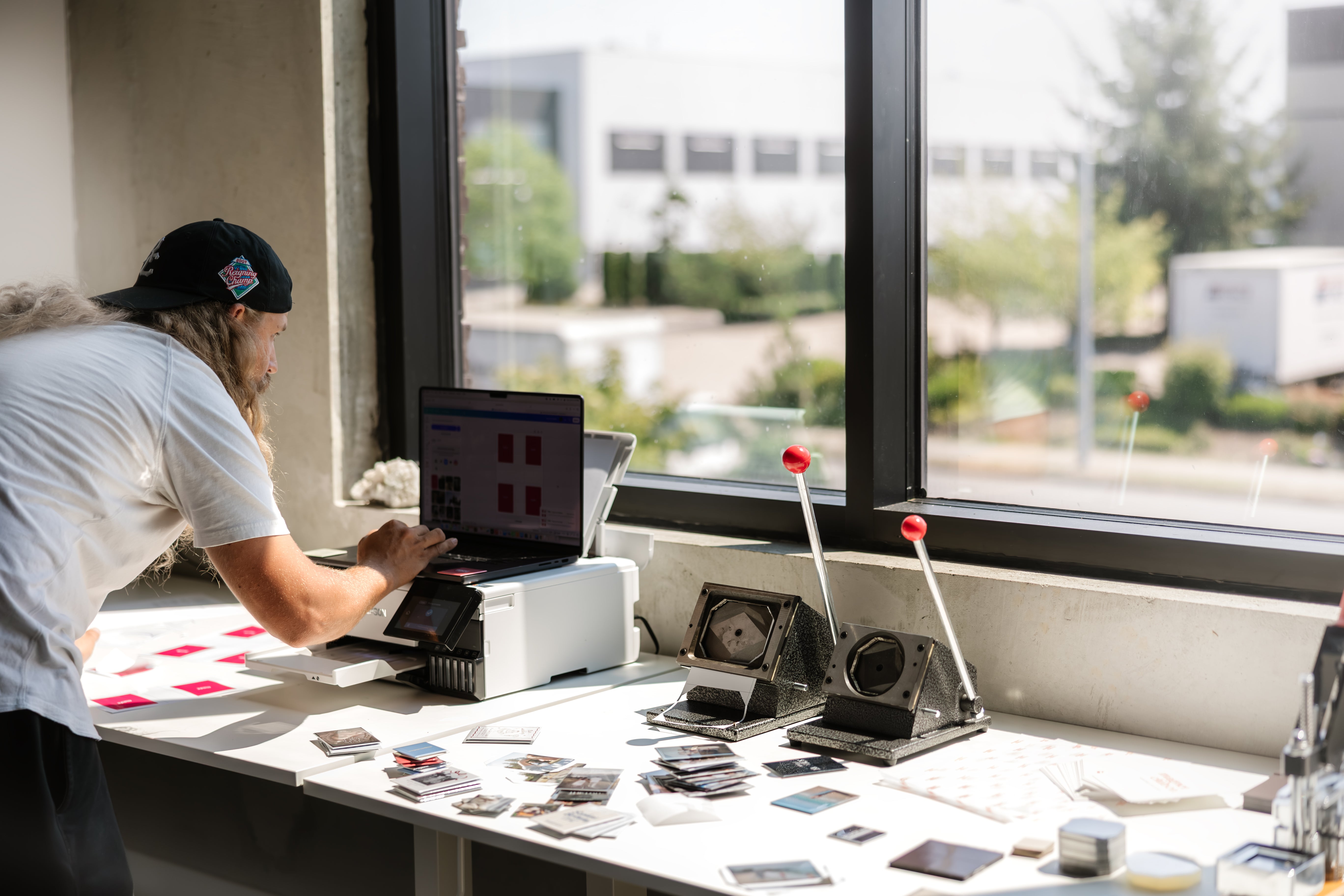 A man in a white t-shirt and black cap works at a table by a large window, using a white desktop computer. The table is covered with small cards, papers, and vintage-style cameras or photo equipment, with an outdoor view visible in the background.