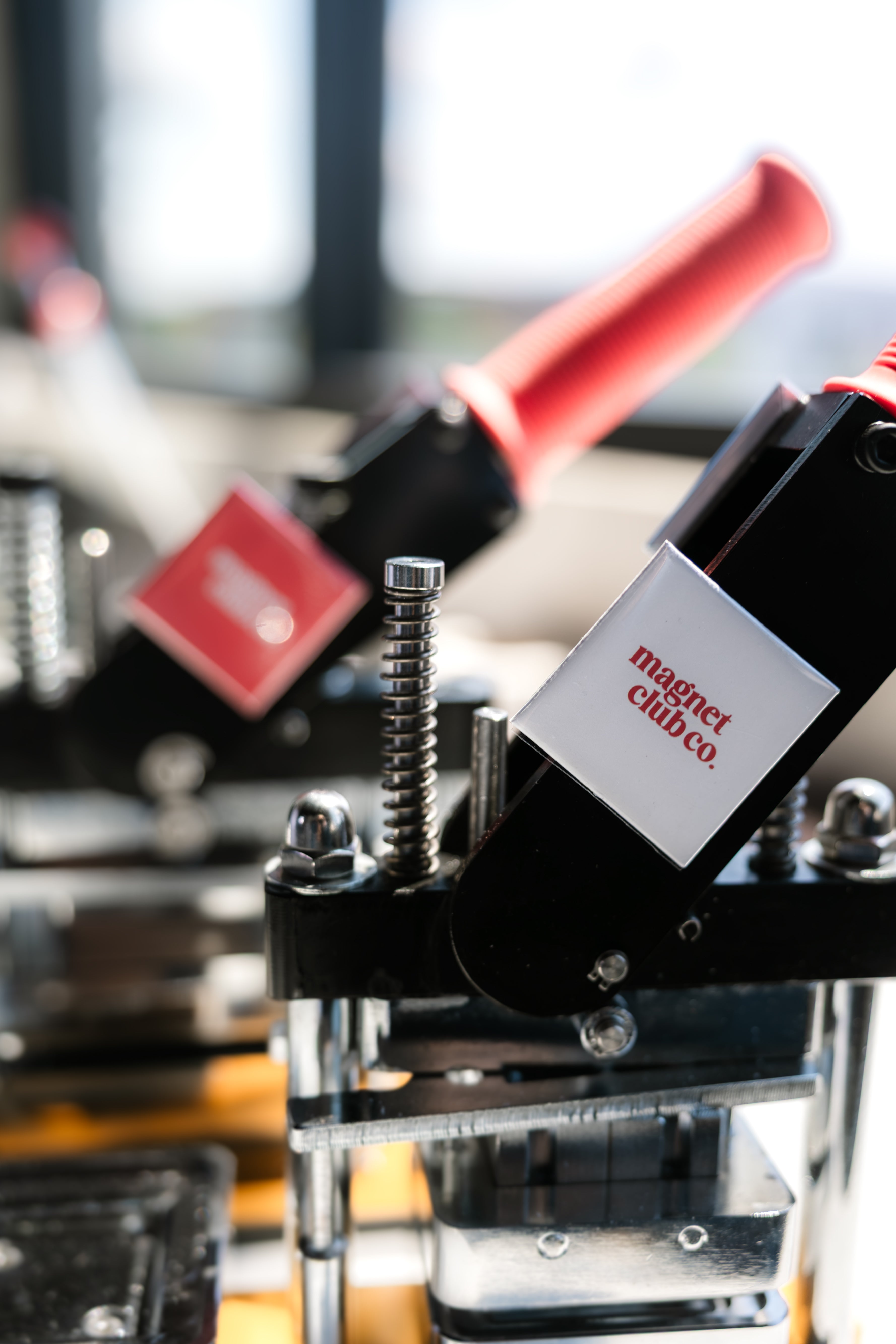 Close-up of a magnetic clamp tool with a black body, silver threaded screw, and a white label reading 'Magnet Club Co.' A red handle is partially visible in the blurred background.